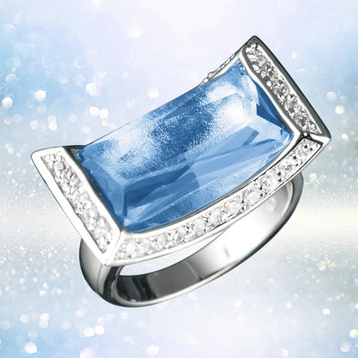 Plateau Ring featuring a rhodium-plated sterling silver band with a large rectangular blue gemstone surrounded by small sparkling clear stones, designed for women’s jewelry.