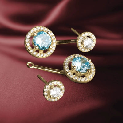 Catalina Halo Earrings featuring sterling silver with sparkling blue and clear gemstones surrounded by a halo of smaller stones, displayed on rich burgundy fabric.