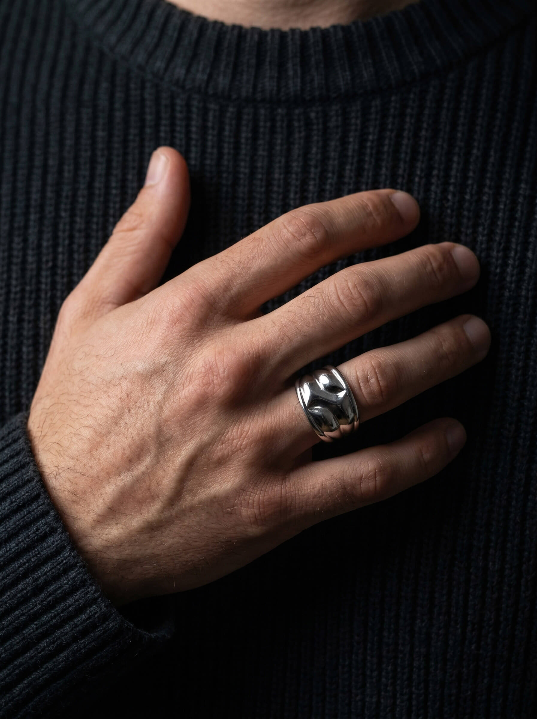 Close-up of a man's hand wearing the Axis Interlock Men’s Ring made of polished silver-tone metal on the ring finger against a dark ribbed sweater background.