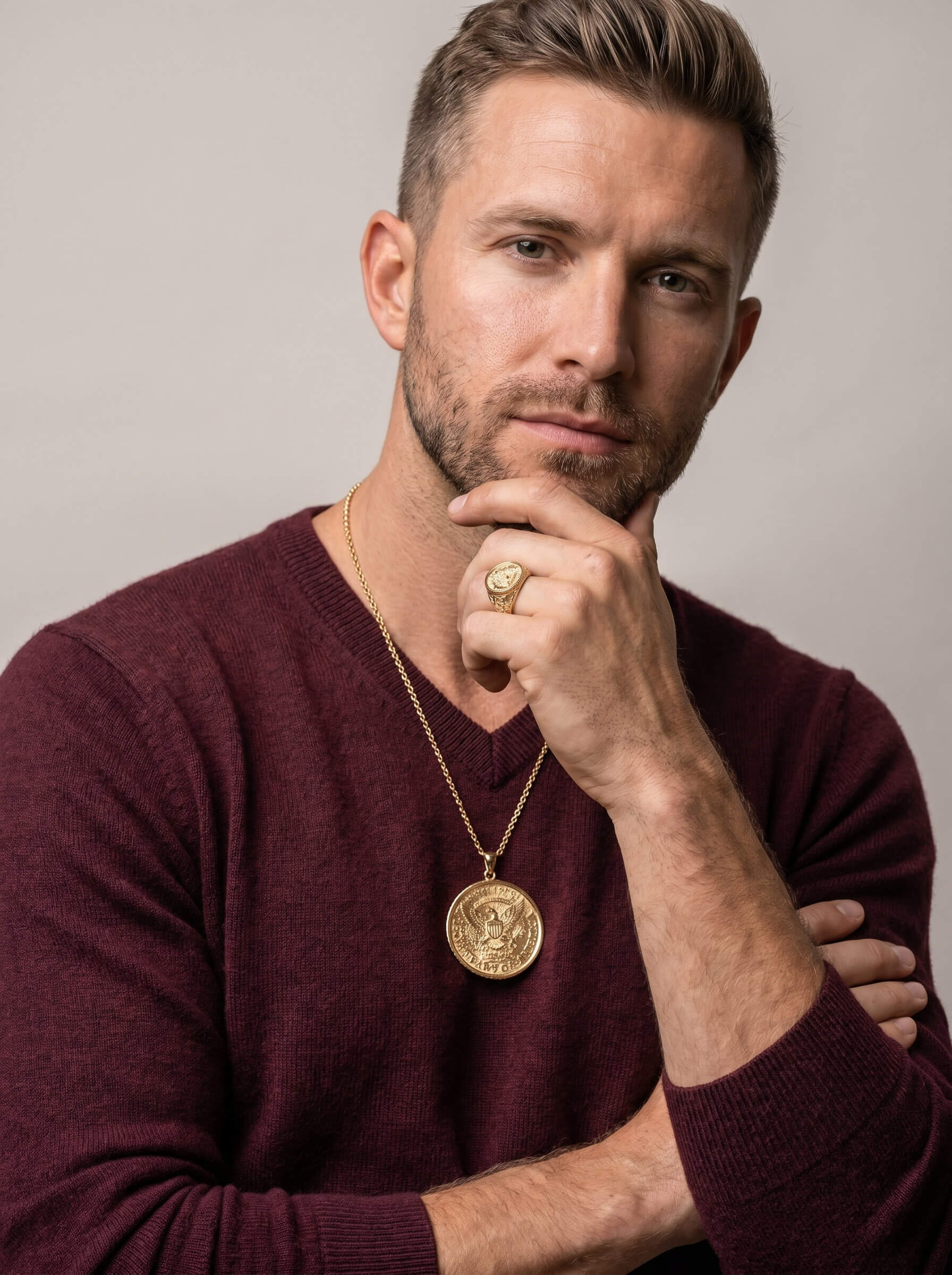 Man wearing a gold American Glory Ring and matching gold pendant necklace dressed in a burgundy sweater against a neutral background