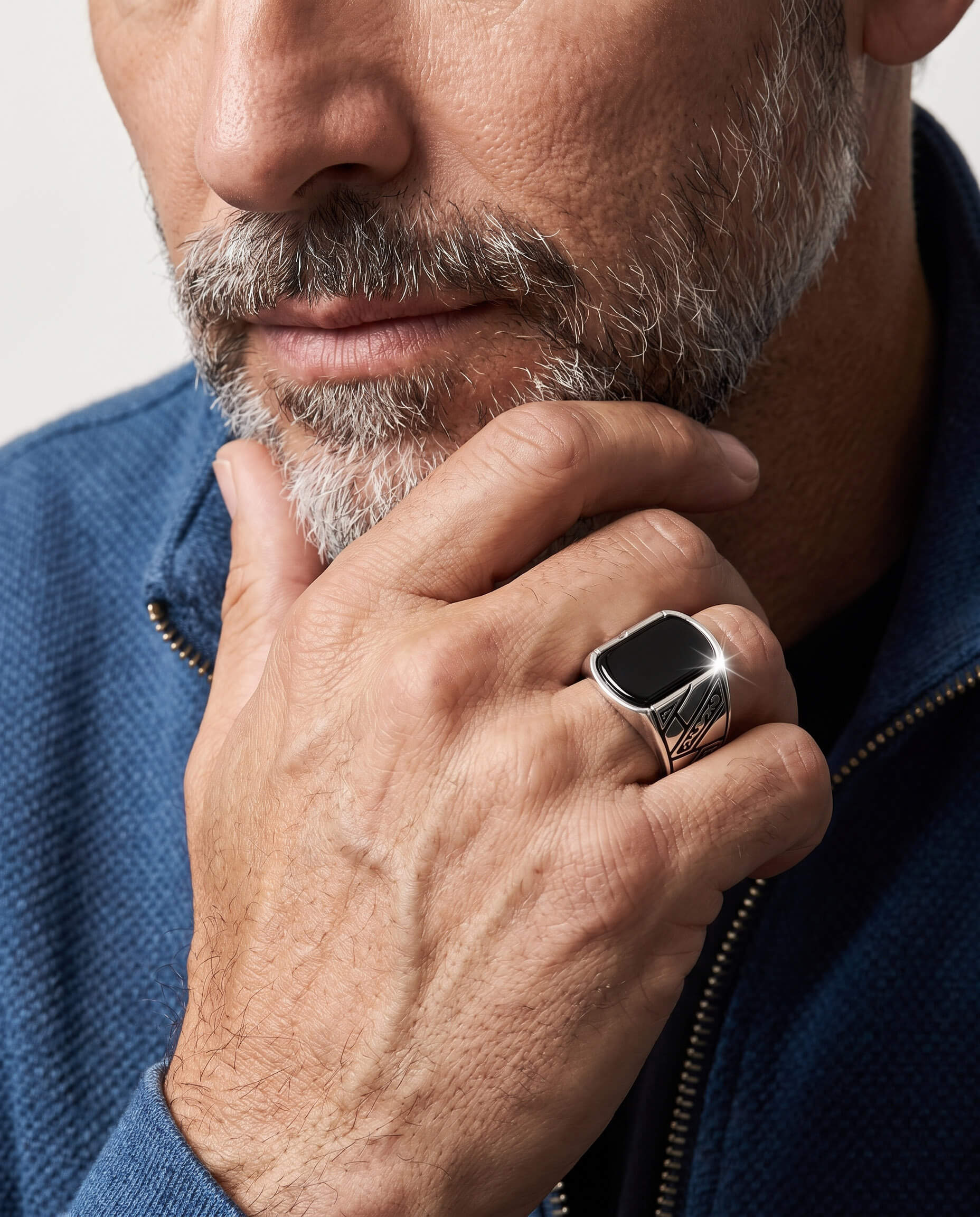 Close-up of a man wearing an Onyx Raven Men's Ring with a polished black onyx stone set in a silver band, showcasing detailed engravings, worn with a blue zip-up sweater, Up To 75% Off Retail.