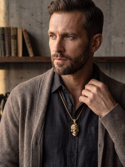 The Fallen Pendant sculpted gold necklace worn over a dark shirt by a bearded man in a textured cardigan with warm indoor styling