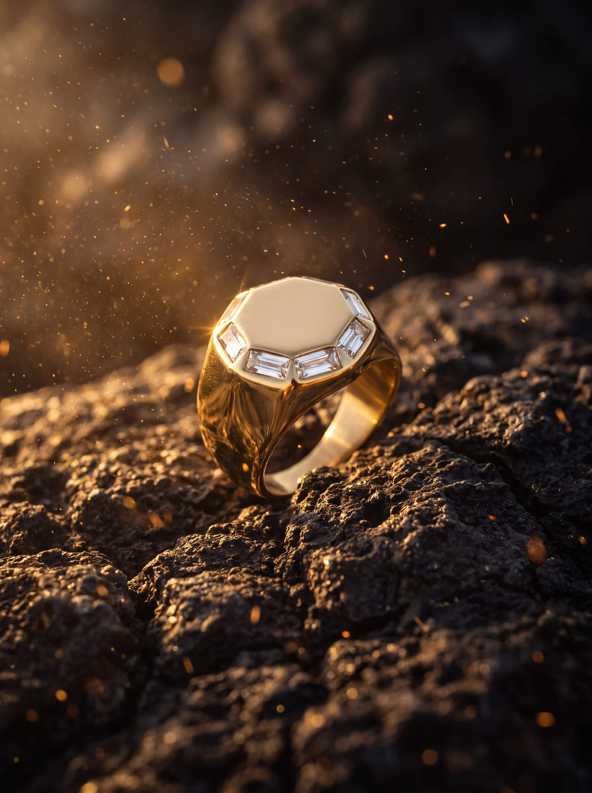 Gold toned signet ring with a faceted octagonal face and clear accent stones resting on rugged dark rock in warm glowing light with floating golden dust