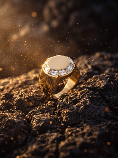Gold toned signet ring with a faceted octagonal face and clear accent stones resting on rugged dark rock in warm glowing light with floating golden dust