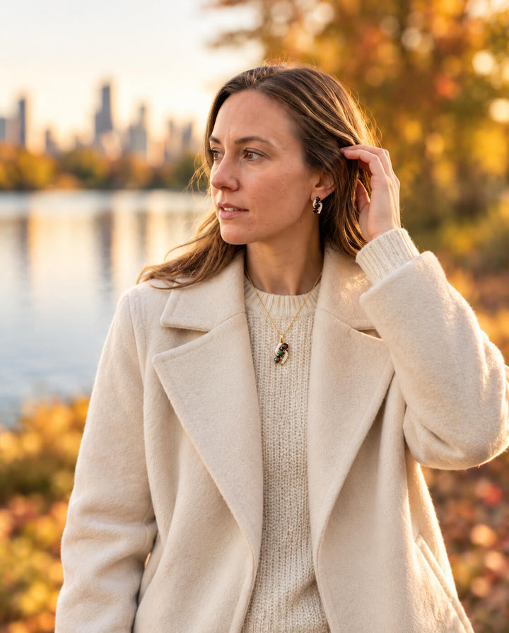 Ember & Eclipse Opal Pendant worn by a woman in a cream coat, featuring an elegant opal design against a sunlit autumn lakeside setting