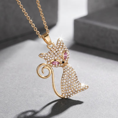 Gold tone cat pendant necklace covered in sparkling clear stones with pink accents and a curled tail displayed against a gray background. Daniel Steiger Crystelle Alley Cat Pendant